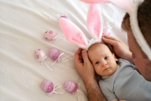 Man wearing funny bunny ears to his newborn son lying on bed among pink eggs. Celebrating Easter